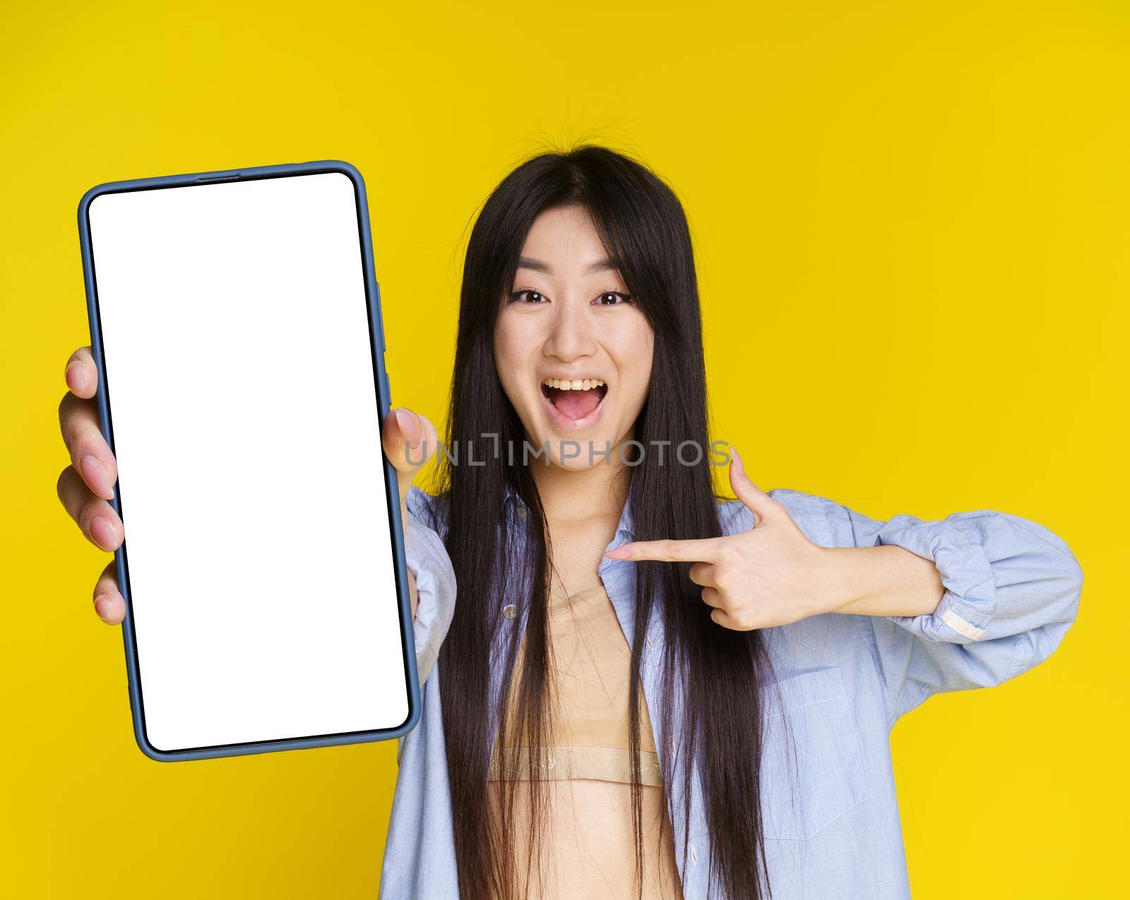 Asian girl excited win, lottery, casino, game, sale offer holding smartphone pointing on empty screen isolated on yellow background. Product placement mock up. Mobile application advertisement.