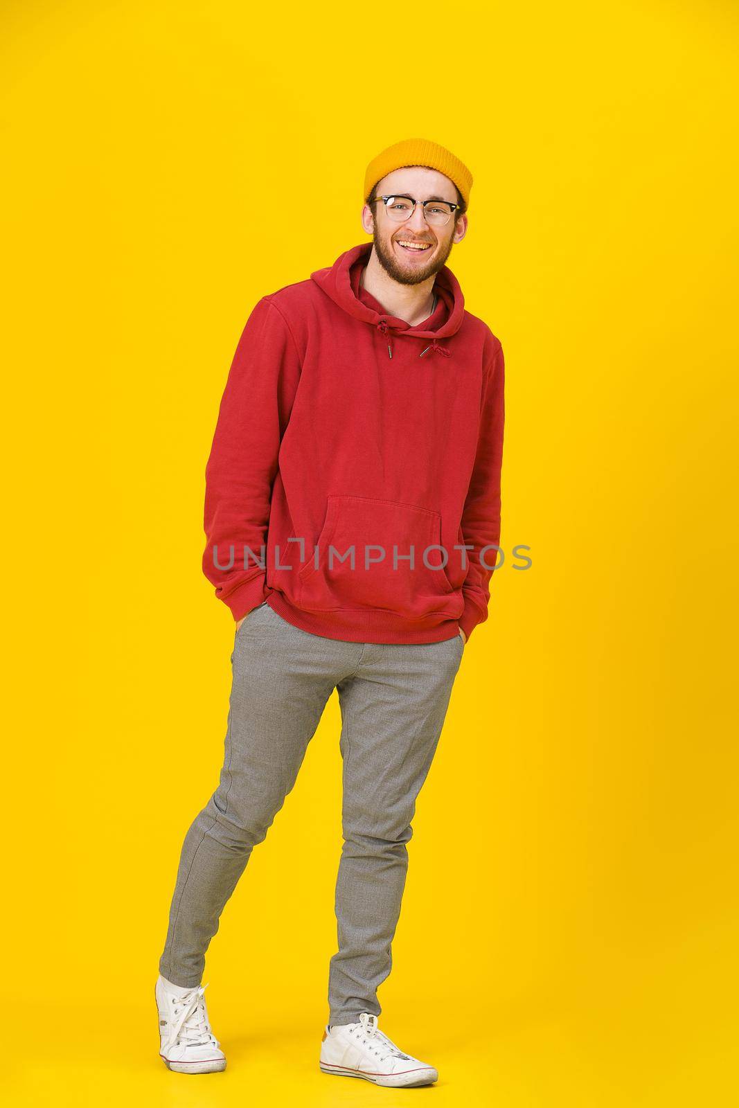 Full body handsome young man in casual wear, glasses with hands in pockets posing looking at camera on yellow background. Hipster fashion bearded smart man casual look.