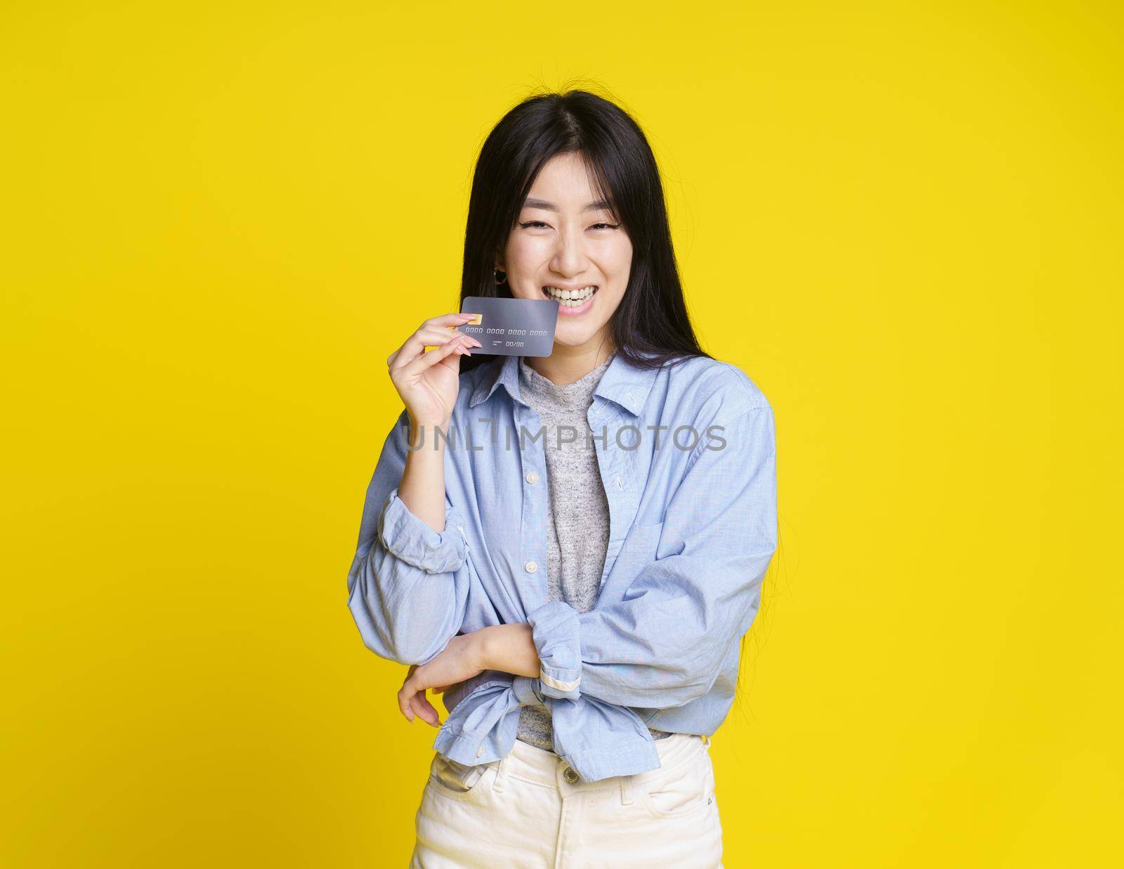 Biting bank card happy asian girl with credit, debit card. Asian girl in blue shirt holding mockup credit card isolated on yellow background. E-banking concept. E-commerce concept.