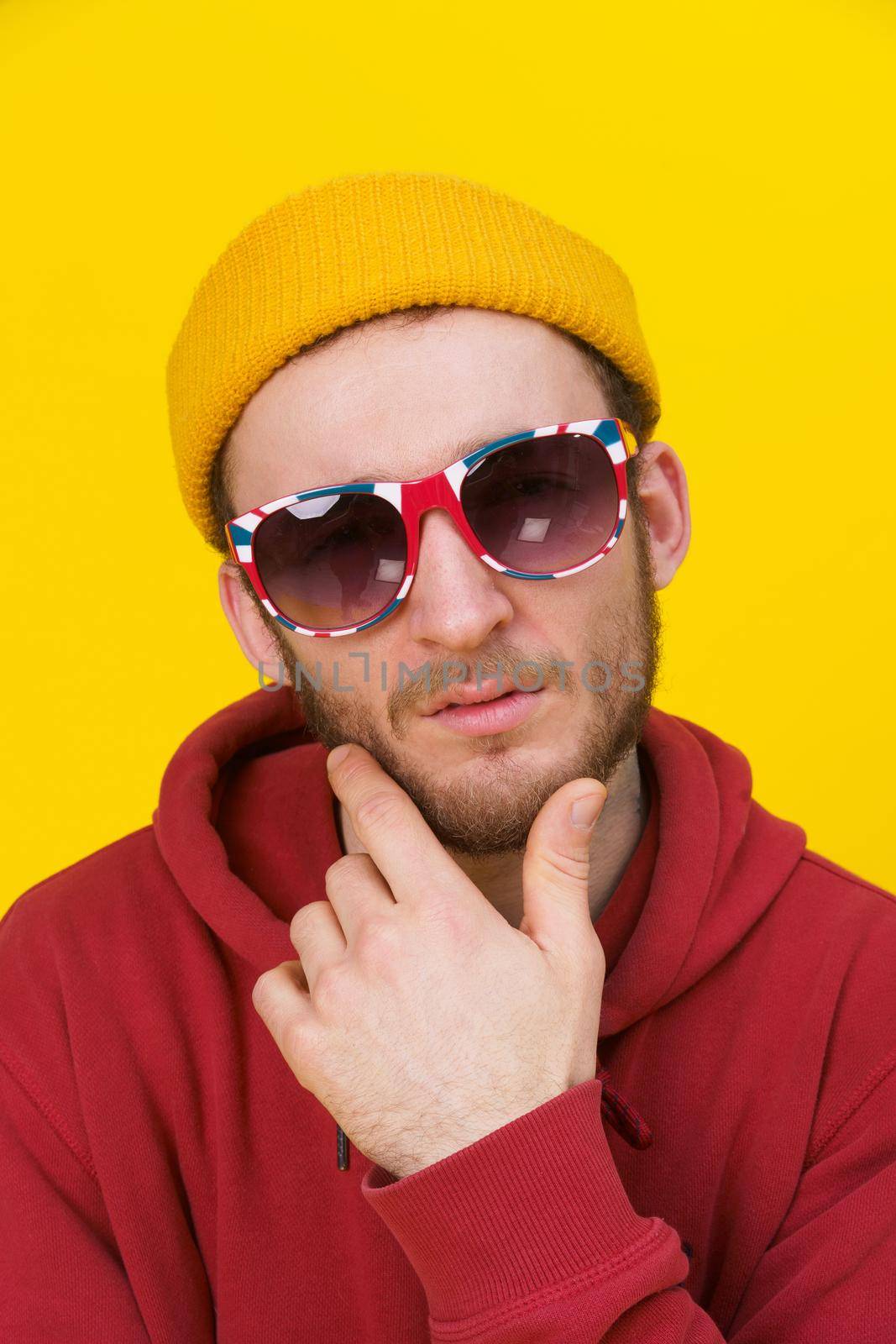 Portrait of a young handsome caucasian man in casual wear, sunglasses looking at camera on yellow background. Stylish bearded smart hipster man casual look. Thoughtful guy on yellow background by LipikStockMedia