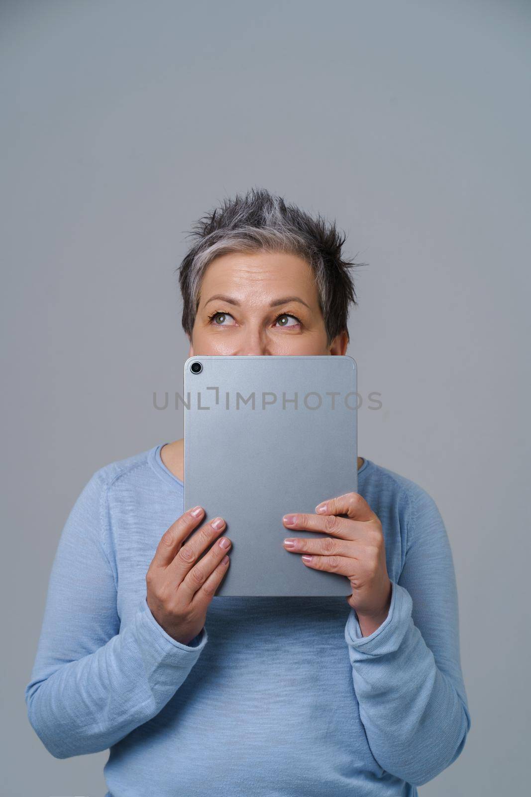 Thoughtful grey haired mature woman thinking hide face behind digital tablet working ideas. Pretty woman with tablet in blue shirt isolated on white background by LipikStockMedia