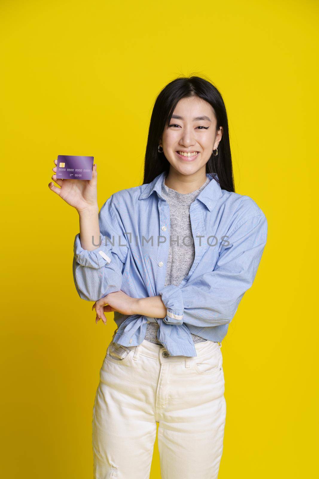 Smiling asian girl holding credit, debit card in hand. Asian girl in blue shirt with mockup banking card isolated on yellow background. E-banking concept. E-commerce concept by LipikStockMedia