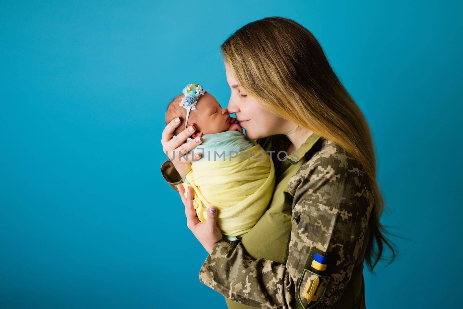 A Ukrainian mother in military uniform with an infant in the colors of a yellow blue flag during the 2022 war. Close-up in the studio, insulated background. The most important heroes of women.