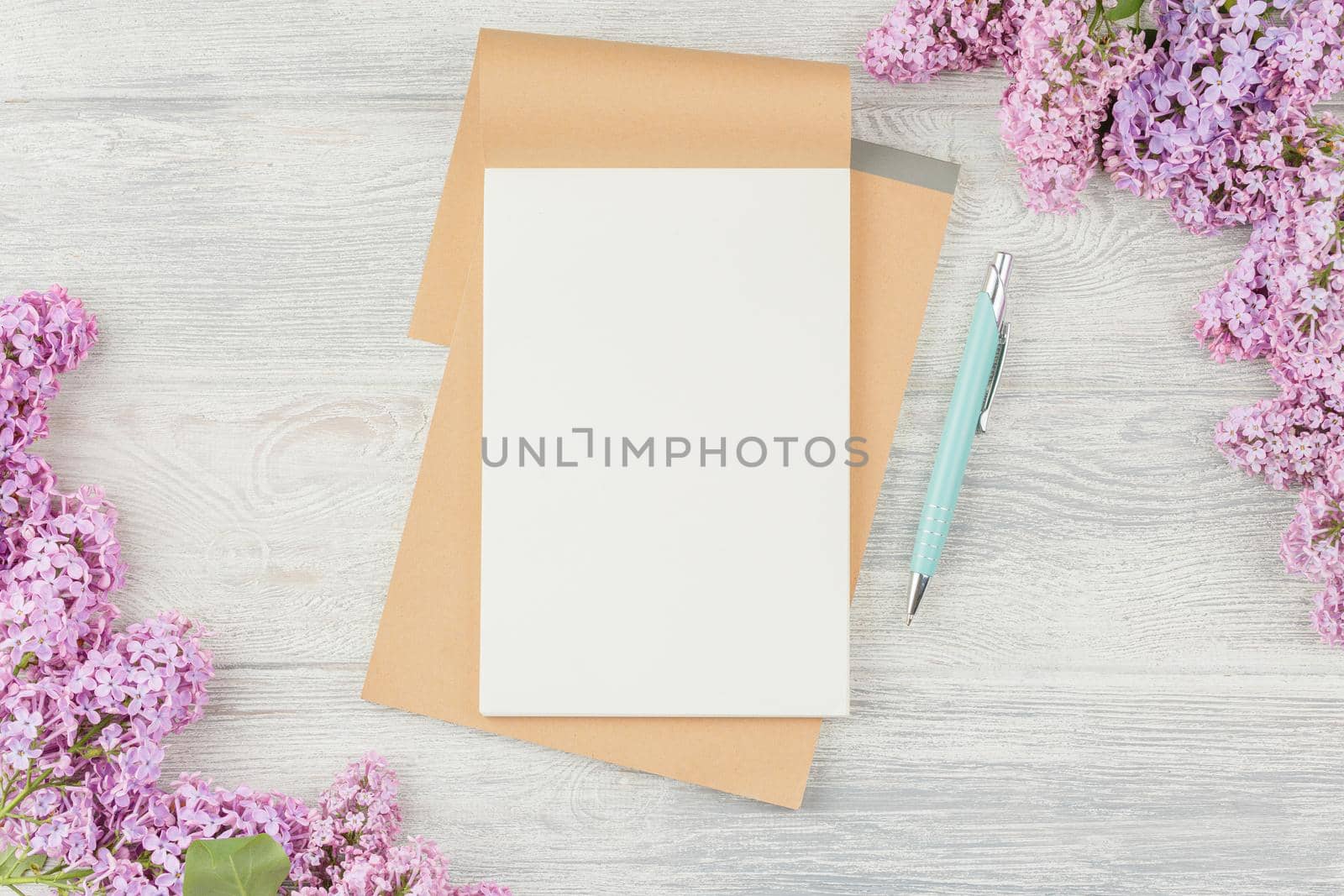 Open craft notebook with pen and lilac flowers on a white wooden background. Top view.