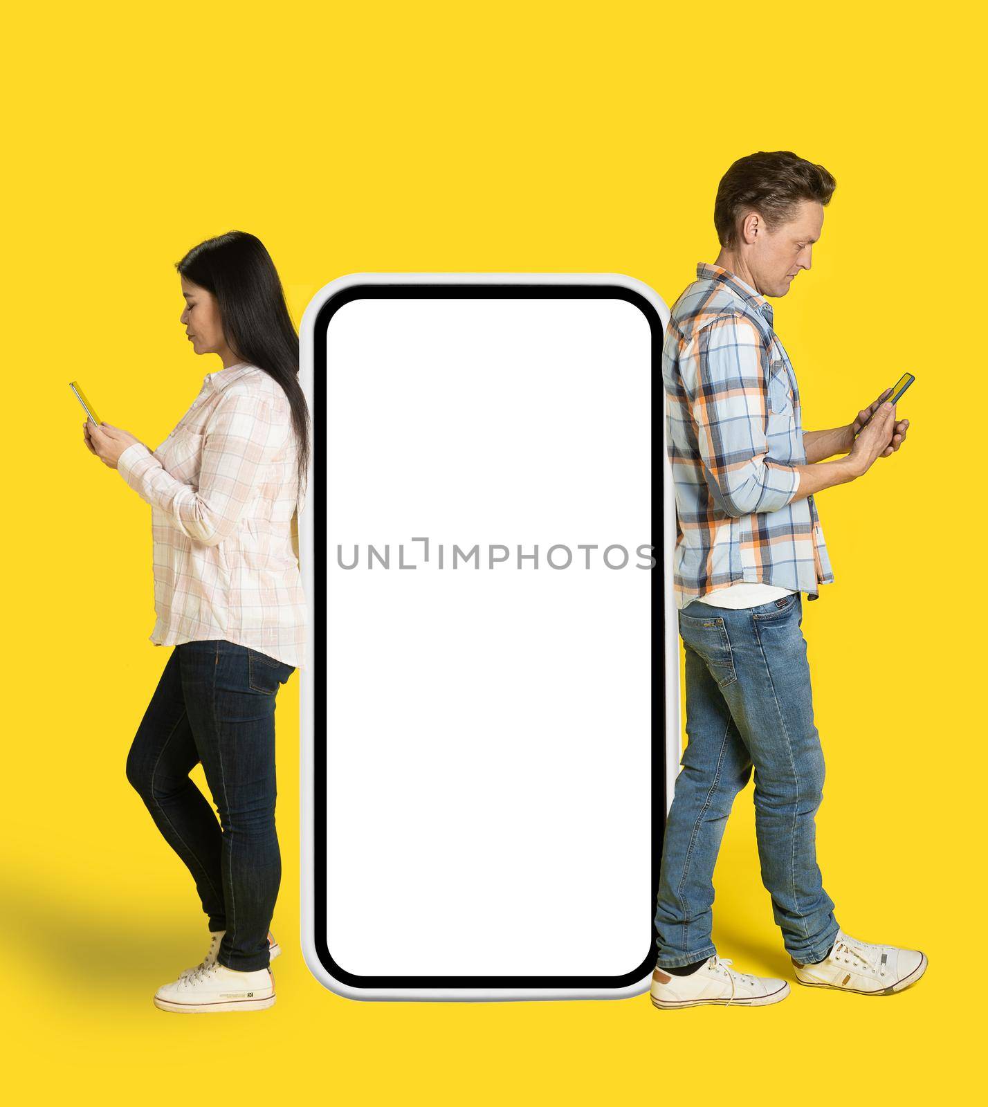 Asian woman and caucasian man standing back to back leaned on huge smartphone with white screen looking at phones in hands, mobile app advertisement isolated on yellow background. Product placement.