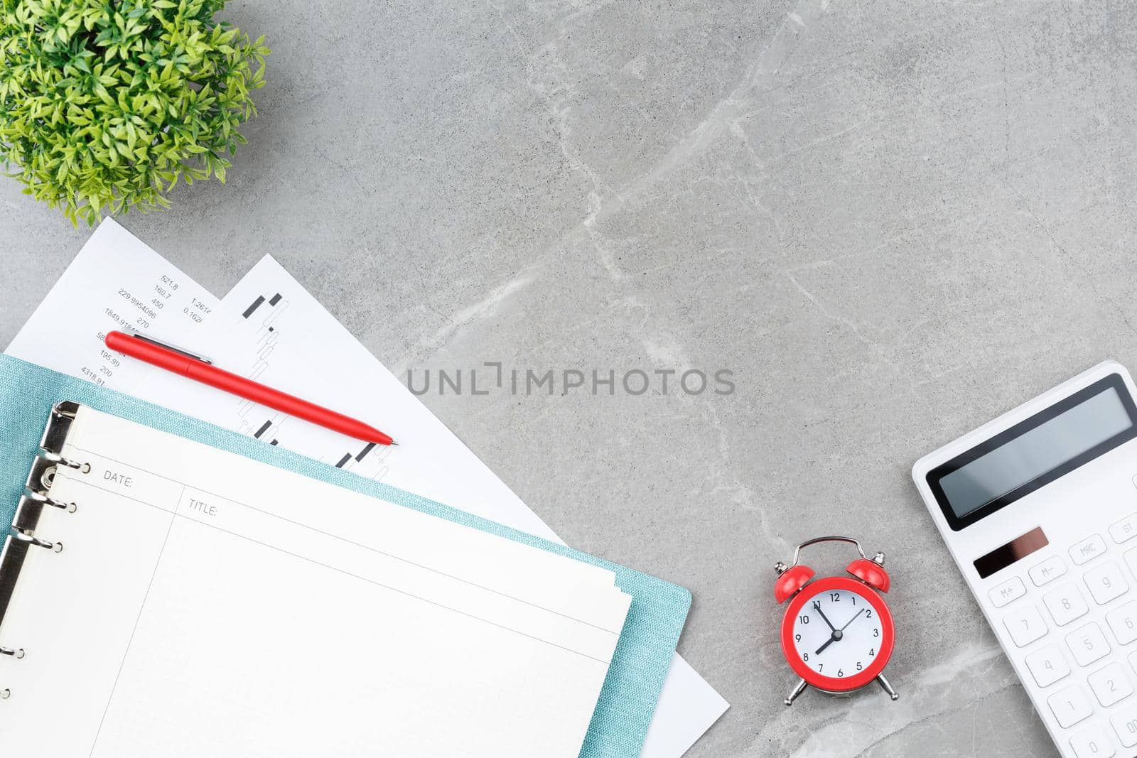 Spiral notebook in soft blue cover with a metal pen, calculator, alarm clock, stock charts and a flower in a pot on a gray marble background. Office desk concept. Top view.