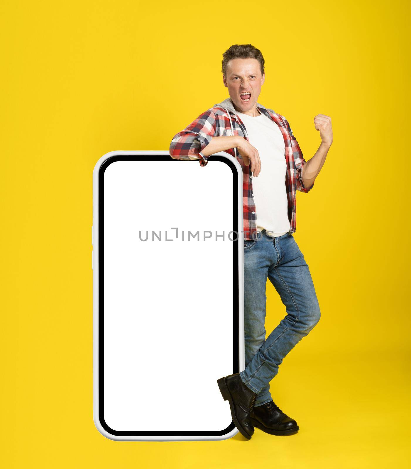 Pointing on huge, giant smartphone with white screen young man leaned on it gesturing with another hand dressed in casual isolated on yellow background. Free space mock up by LipikStockMedia