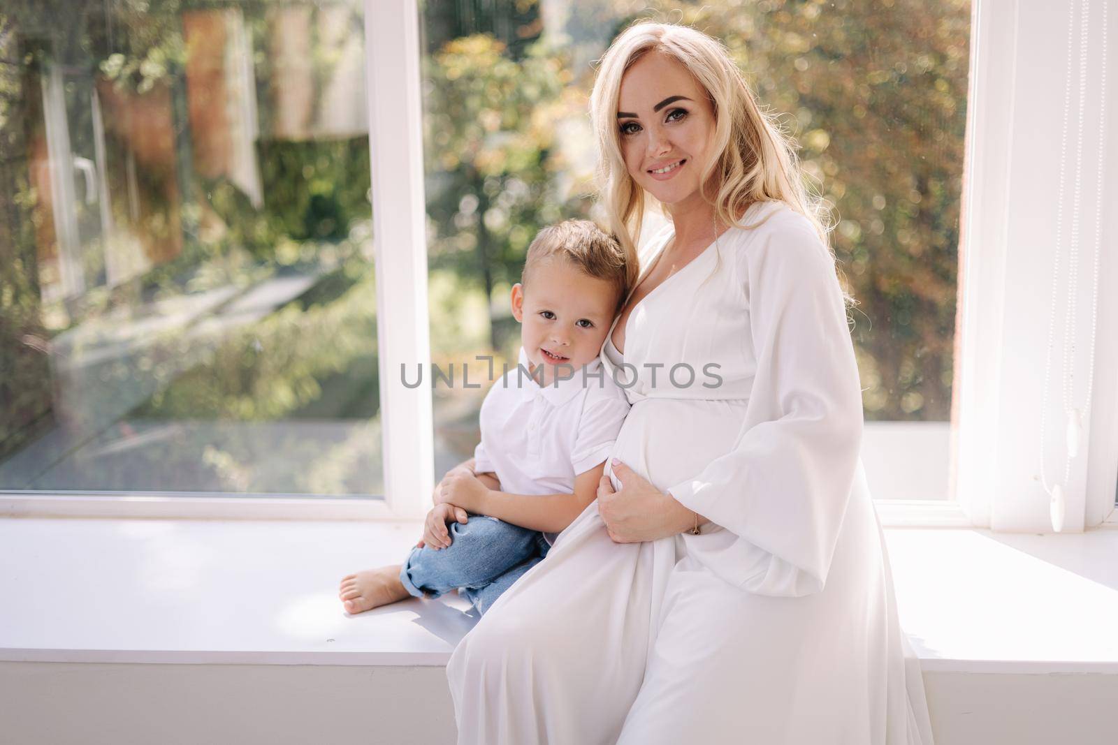 Beautiful pregnant woman with her son. Boy hug his mom and rejoices. Boy waiting for sister. Lady in elegant white dress posing to photographer in studio. Background of white tulle.