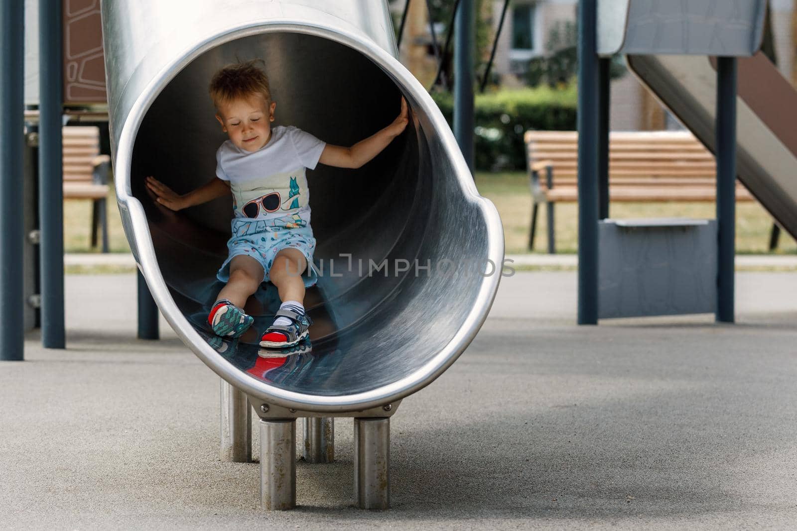 Boy in a white t-shirt blonde, riding on a metal slide tube on the playground.