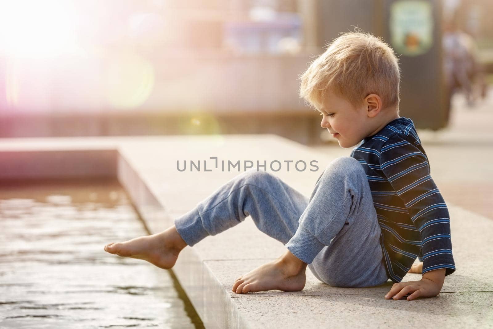 Cute little boy playing with water hot summer outdoors, the child tests the temperature of the water in the fountain by putting his foot into the water.