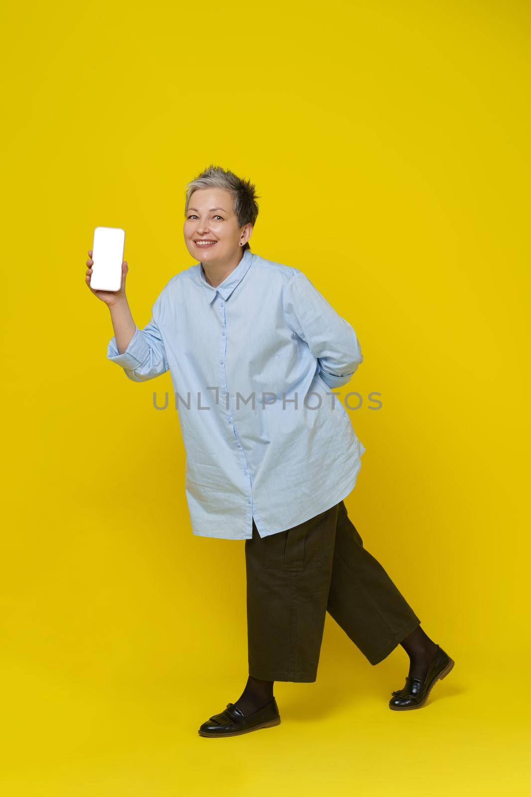 Showing phone in hand mature grey haired woman smiling on camera. Pretty woman in blue shirt and brown trousers isolated on yellow background. Mobile app advertising. Mockup product placement by LipikStockMedia