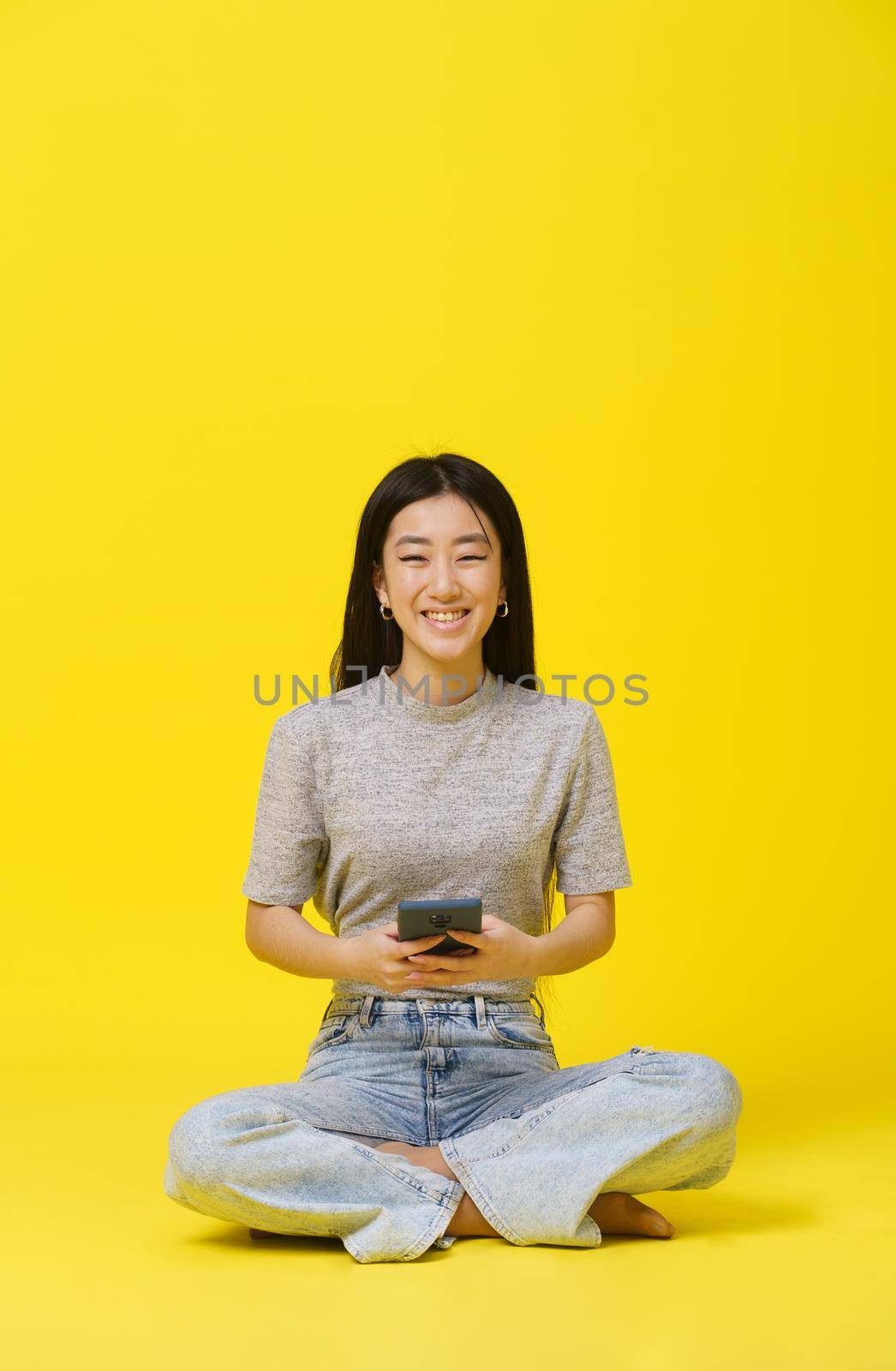 Tender asian young girl sitting on the floor with phone in hands texting or shopping online playing game isolated on yellow background. Mobile App Advertisement. Copy space by LipikStockMedia