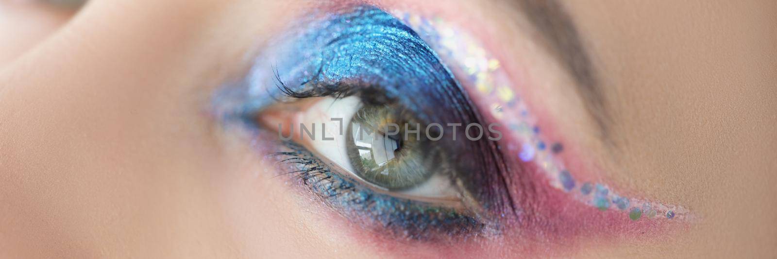 Close-up of festive bright eye makeup on womans face with blue and pink glitter. Cats eye technique and colourful mix of eyeshadows. Mua, creative concept