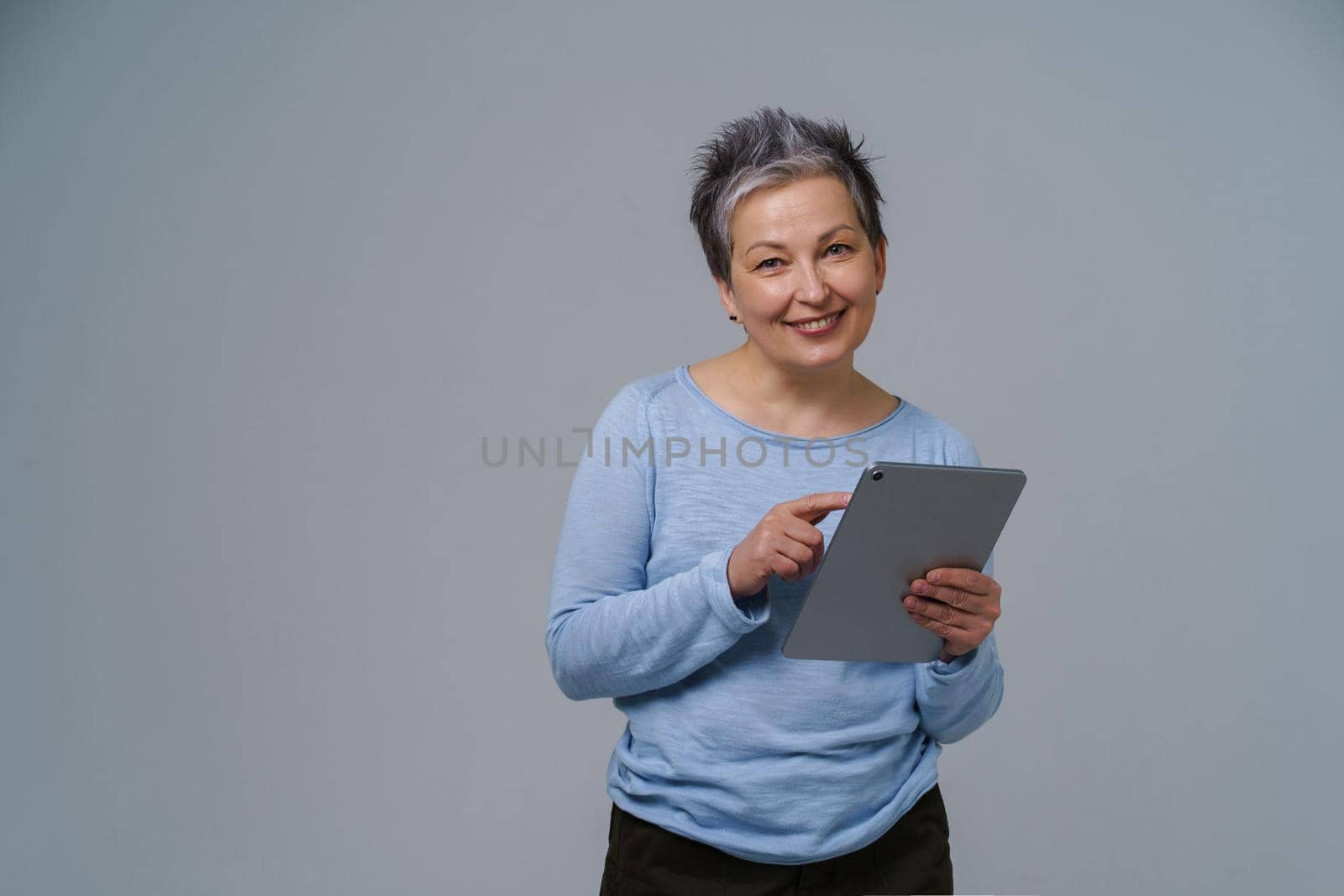 Mature grey haired businesswoman with digital tablet in hands working online. Pretty woman in 50s in blue blouse isolated on white. Older people and technologies.