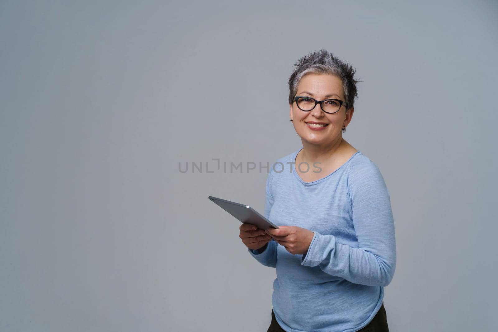 Businesswoman in eye glasses and digital tablet in hands working online smile looking at camera. Pretty woman in 50s in blue blouse isolated on white. Older people and technologies.