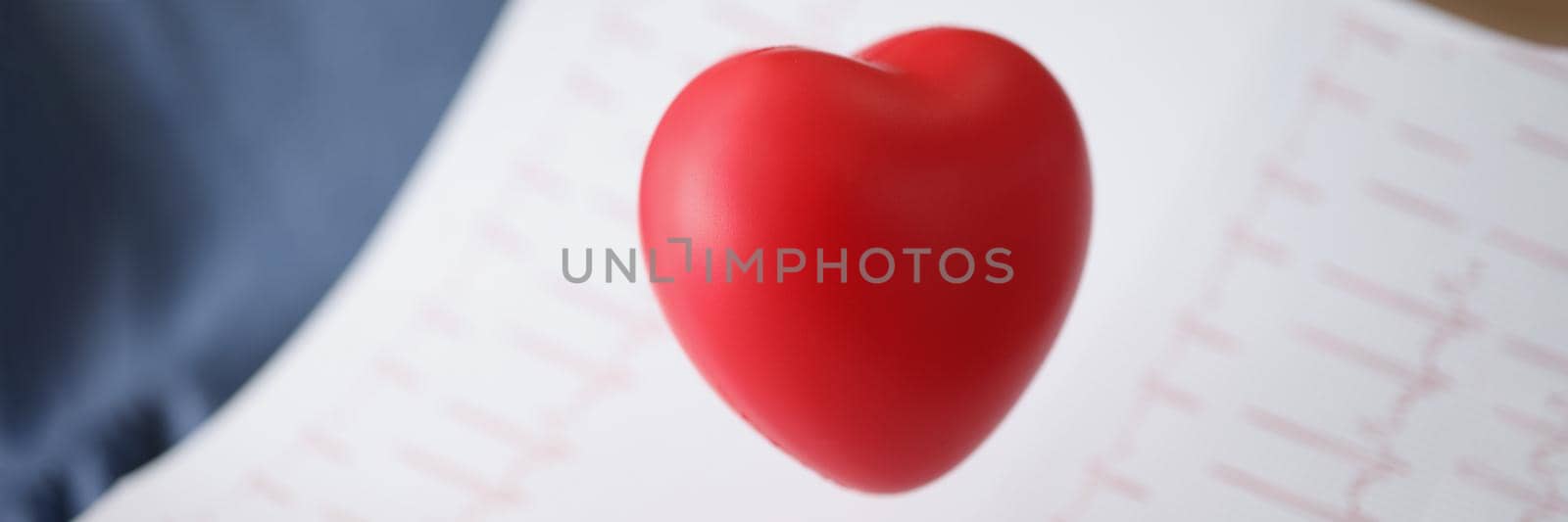 Close-up of red plastic heart and paper with electrogram result. Planned checkup at clinic, heart diagnostic. Ecg, cardiology, healthcare, medicine concept