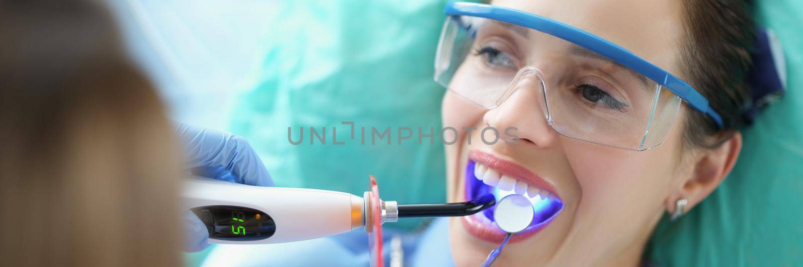 Portrait of woman client in stomatology clinic get teeth whitening with special equipment. Patient wear protective glasses, open mouth. Teethcare concept