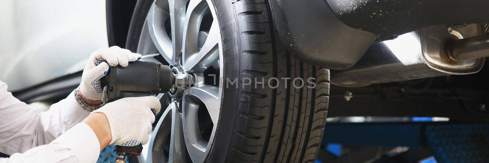 Close-up of automechanic with electric screwdriver changing tire in garage. Handyman worker fix machine on station. Car maintenance, repair service concept