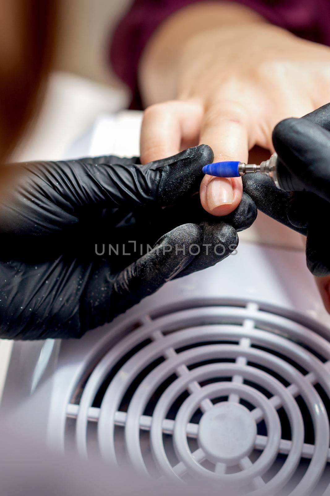 Manicure master applying electric nail file drill to fingernails of young woman in nail salon