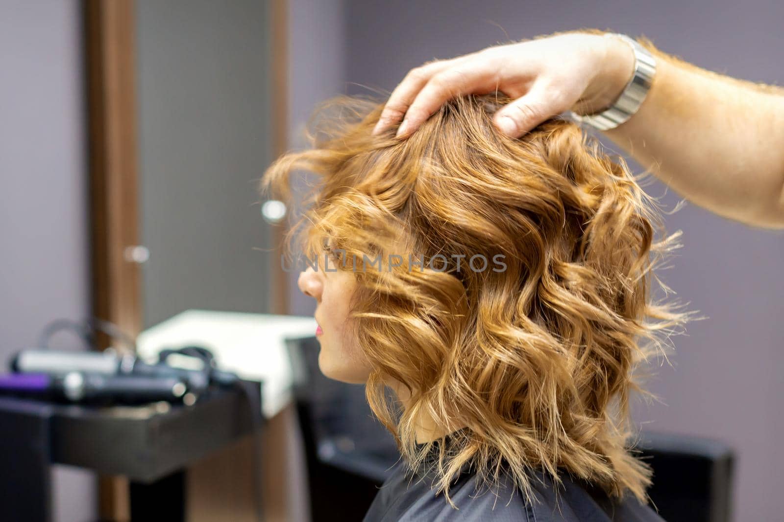 Side view of male hairdresser checks curly hairstyle of young caucasian woman in hair salon. Soft focus