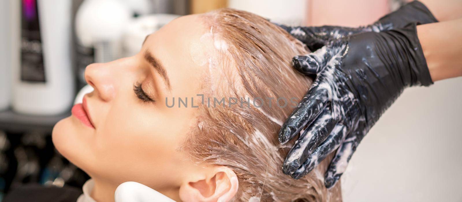 Side view close up of beautiful young caucasian woman receiving hair wash by hands of hairstylist in hair salon. Selective focus