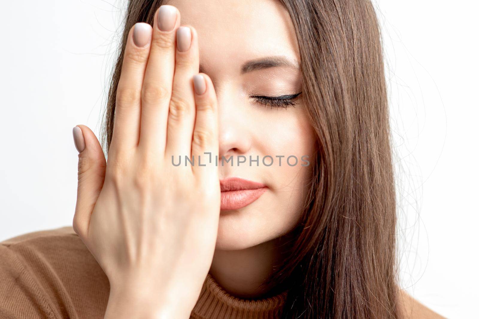 Portrait of beautiful young woman with closed eyes covering one eye by her hand on white background