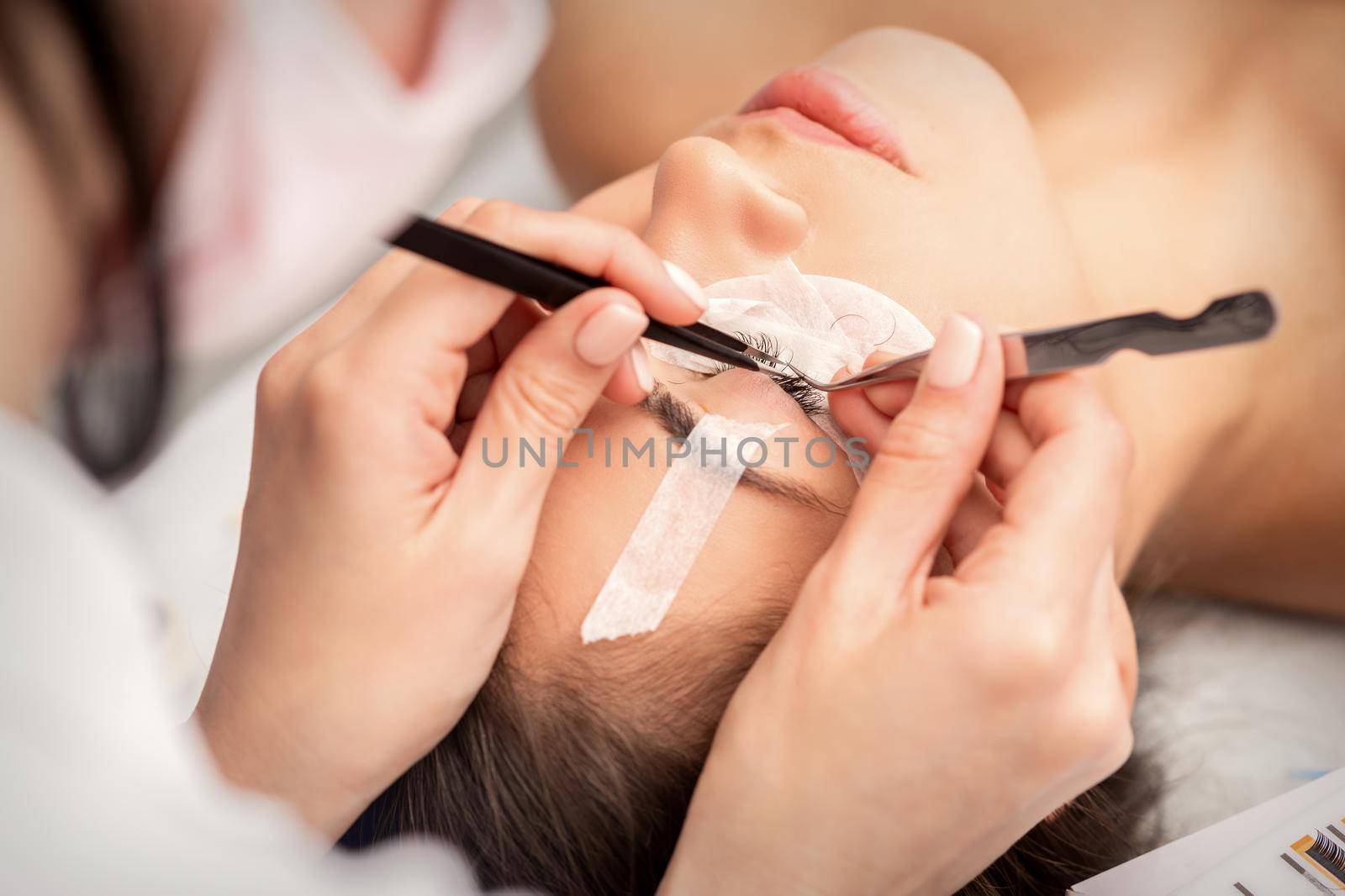 Young caucasian woman having eyelash extension procedure in beauty salon. Beautician glues eyelashes with tweezers
