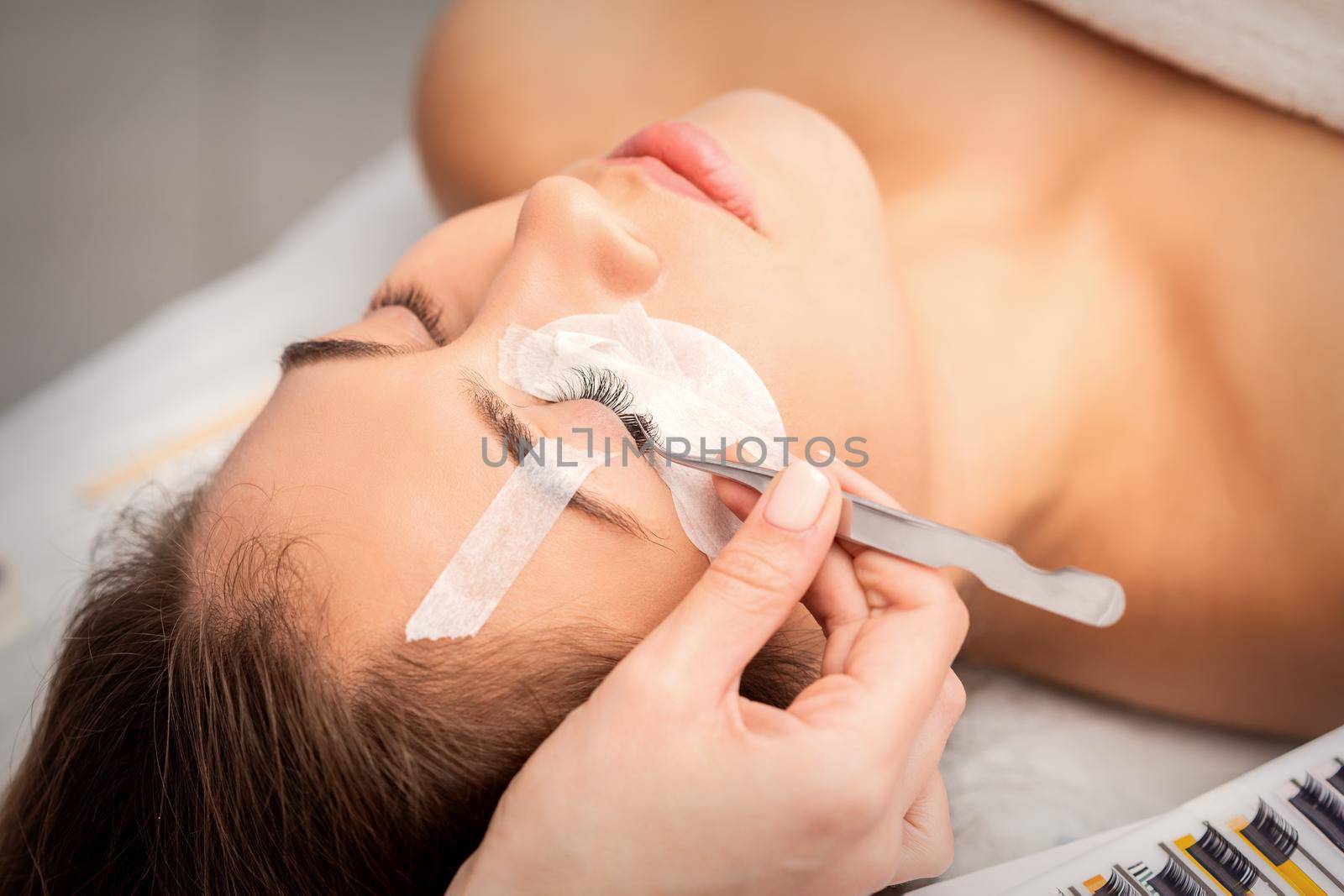Young caucasian woman having eyelash extension procedure in beauty salon. Beautician glues eyelashes with tweezers