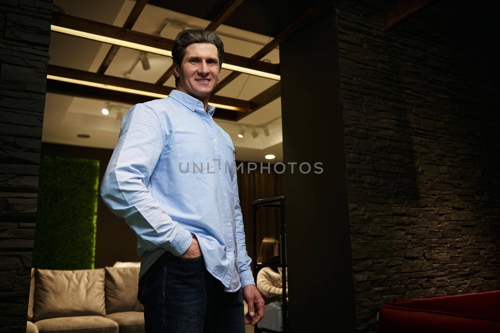 Confident mature Caucasian man - entrepreneur, sales assistant in casual blue shirt and blue denim jeans smiling looking at camera standing in cozy stylish furniture store interior exhibition center