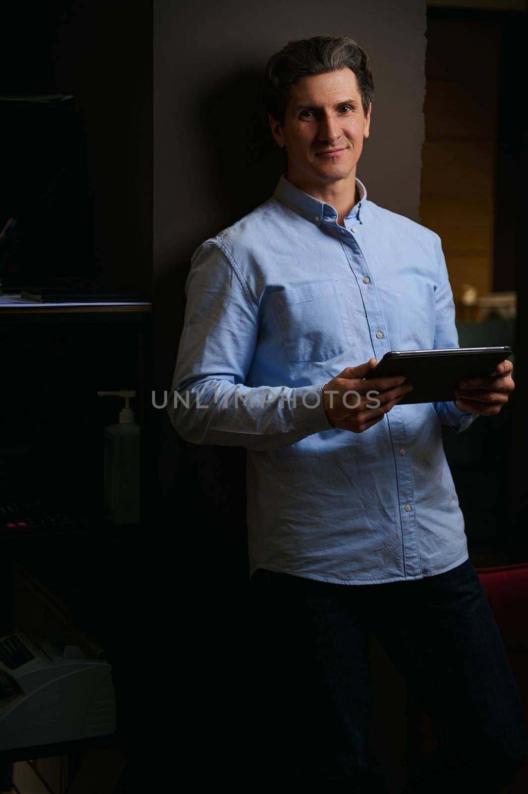 Portrait of confident businessman, successful entrepreneur, real estate agent, fashion designer, creative person with tablet posing in low light office, looking at camera by artgf