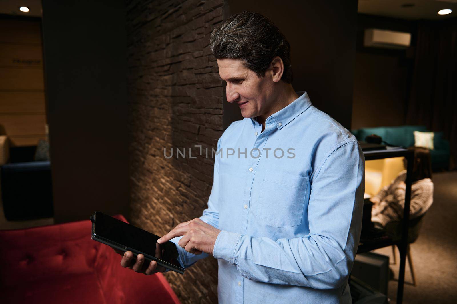 Confident furniture store retail assistant, prosperous sales manager pointing on a blank screen of a digital tablet with copy ad space, standing in a stylish modern interior of a showroom office