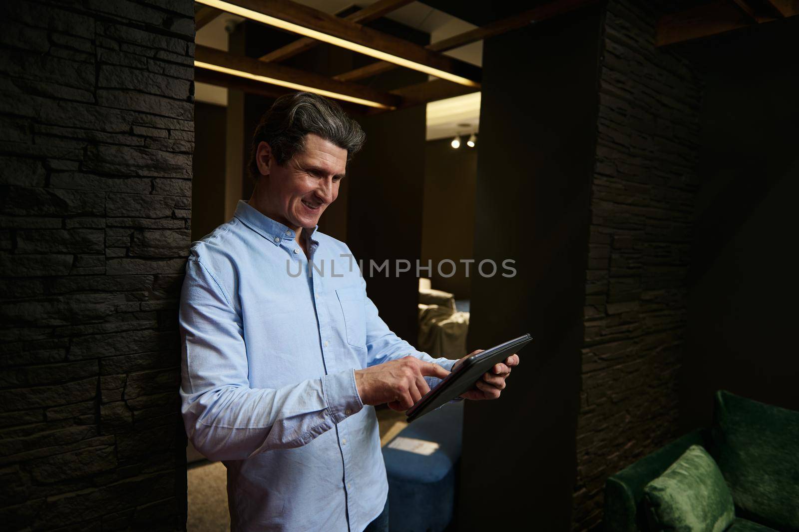 Confident prosperous sales man, real estate agent, interior designer working at a furniture store, using digital tablet and checking orders of new upholstered furniture in an interior design showroom