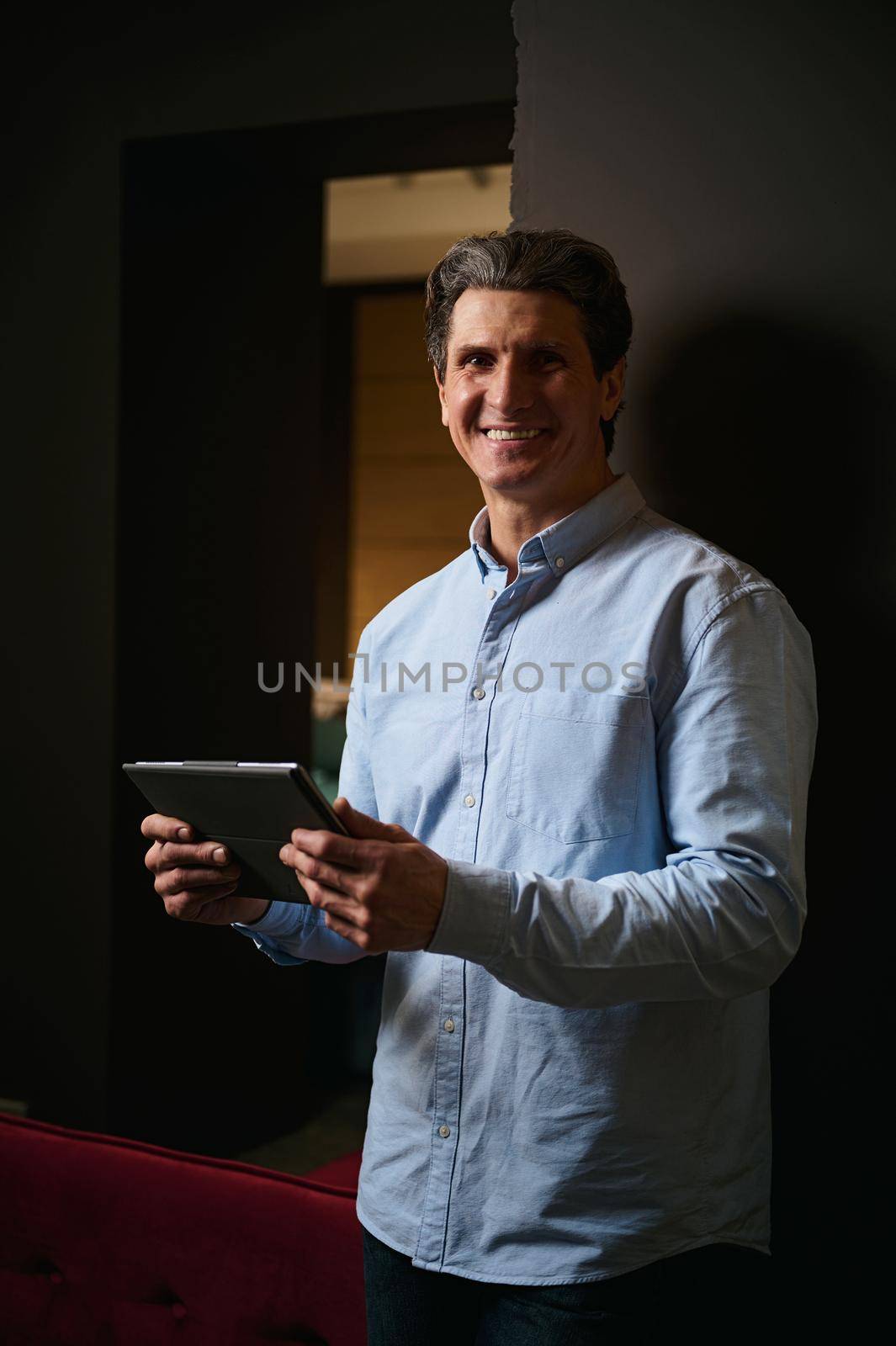 Handsome successful mature Caucasian man holds digital gadget and smiles looking at camera. Interior designer, real estate agent, entrepreneur in a furniture design showroom.