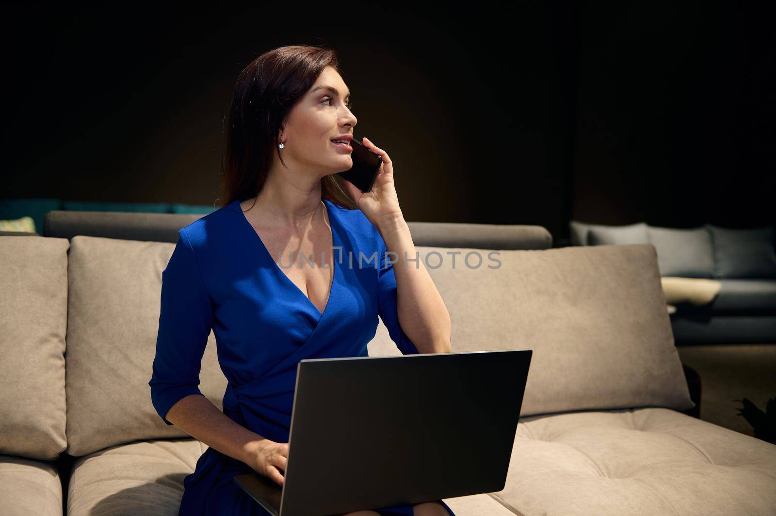 Attractive confident dark-haired Caucasian woman, sales manager, interior designer talks on mobile phone and works on laptop, sitting on a sofa in an exhibition center of furniture design studio