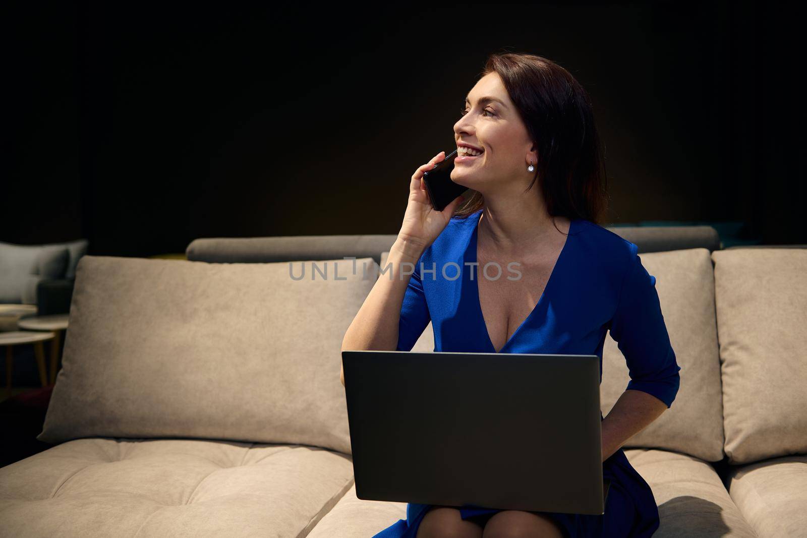Confident business woman, professional interior designer in elegant casual dress talking on mobile phone while working on laptop and planning new home design project by artgf