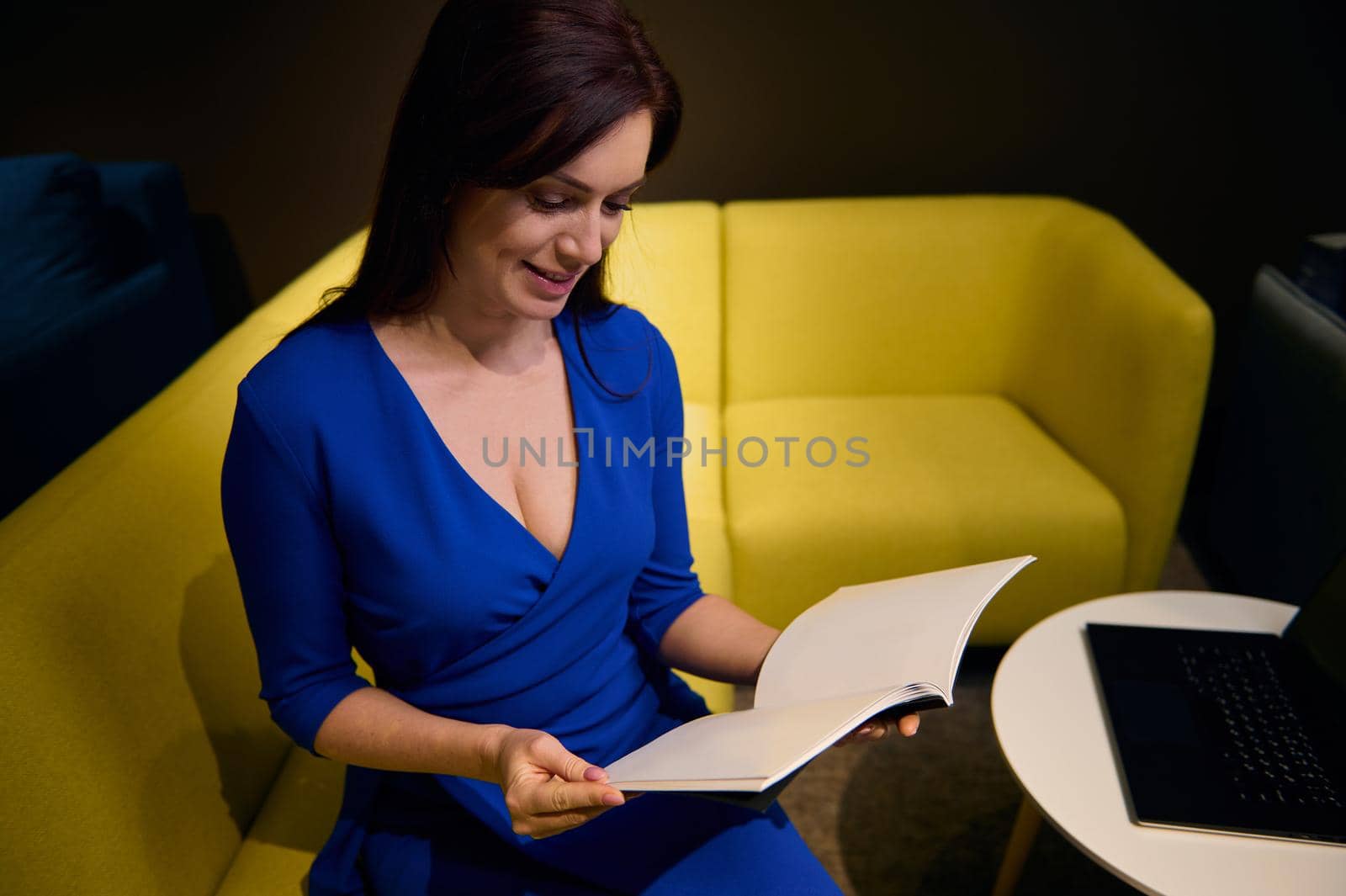 Top view of a beautiful brunette woman leafing through a catalog, sitting on a comfortable yellow sofa next to a coffee table with a laptop and looking for new upholstered furniture in a design studio