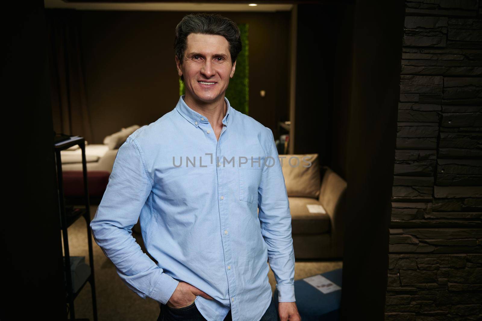 Handsome middle aged caucasian man - businessman, entrepreneur, interior designer, sales manager in casual blue shirt, smiles looking at camera, standing at his workplace in a furniture design store
