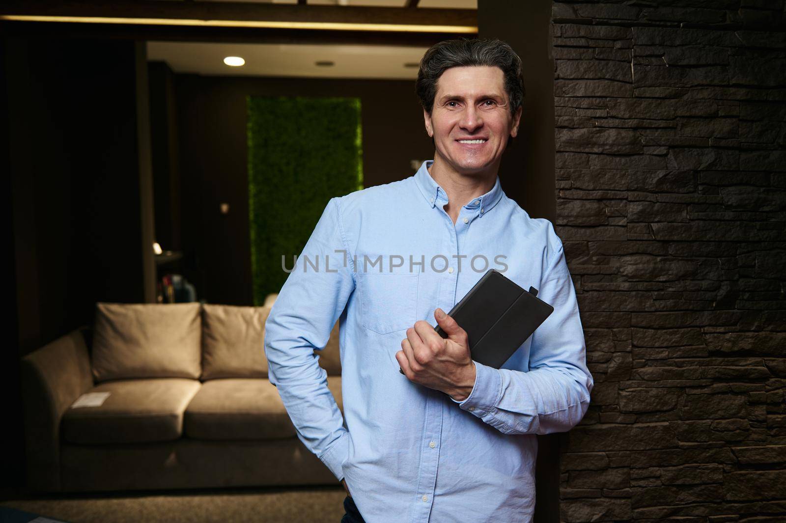 Handsome successful interior designer at home furniture store holding a tablet while smiling at camera. Small business, interior design concept by artgf