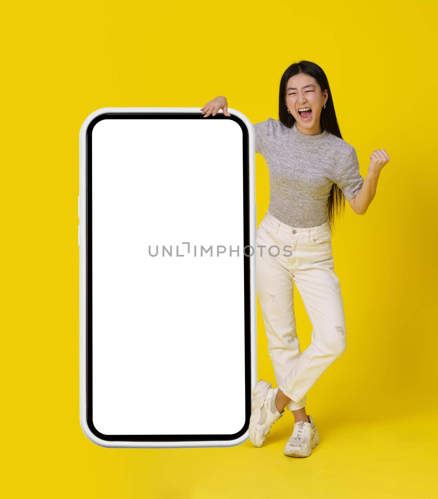 Full body asian girl stand near huge, giant smartphone with white screen happy making yeah, winner hand gesture dressed in casual isolated on yellow background. Free space mock up by LipikStockMedia