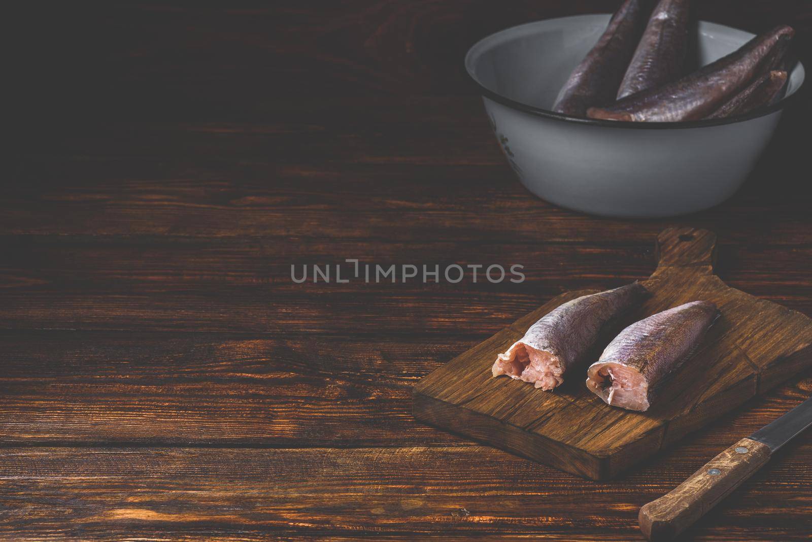 Hake carcasses on cutting board with knife and bowl over wooden surface