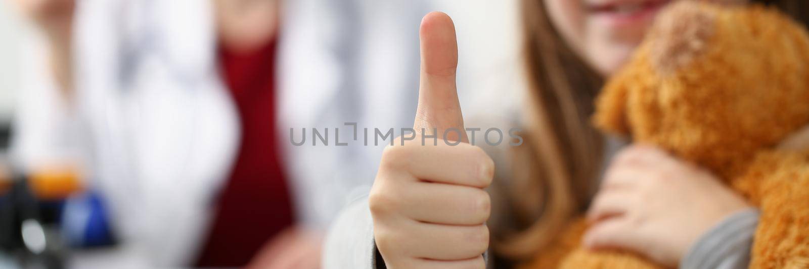 Close-up of smiling child showing thumbs up after appointment in doctor office. Schoolgirl with bear visited family doctor for checkup. Medicine concept