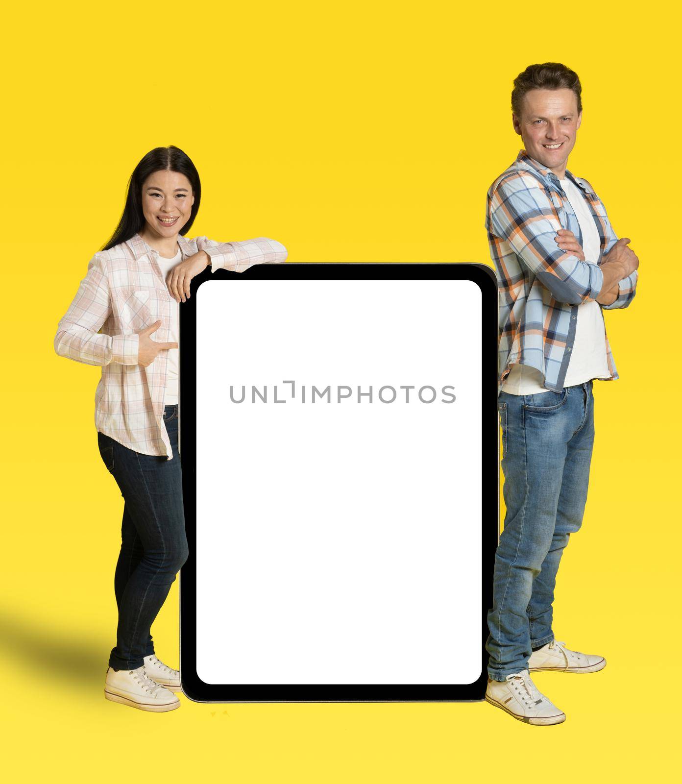 Asian woman and caucasian man back to her leaned on huge tablet pc with white screen, mobile app advertisement looking at camera isolated on yellow background. Product placement.