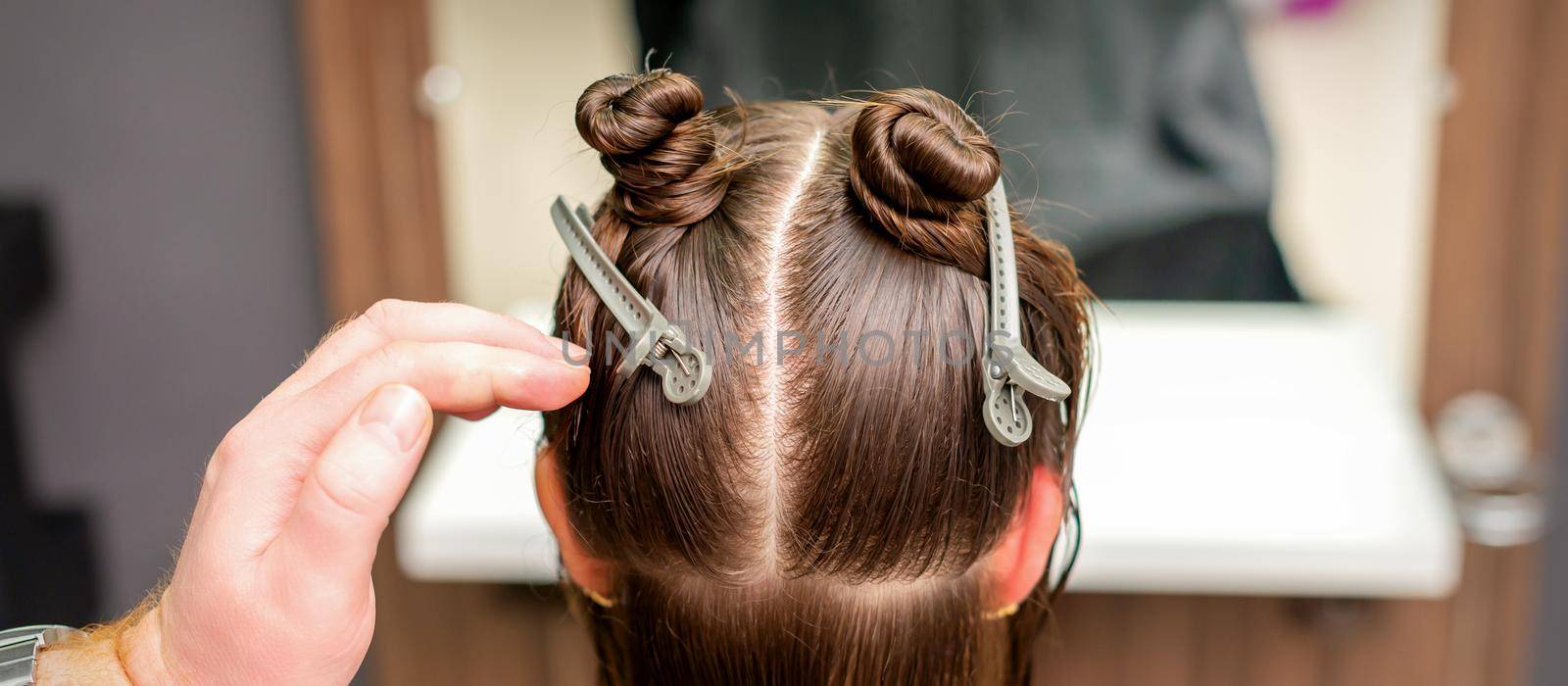 Rear view of hand of hairdresser doing haircut of young woman with hair clips on her hair in hair salon