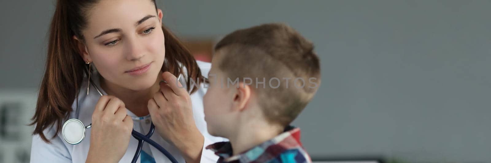 Portrait of little boy sitting on doctor appointment, qualified pediatrician wear stethoscope. Woman doctor in medical gown talk to kid. Medicine concept