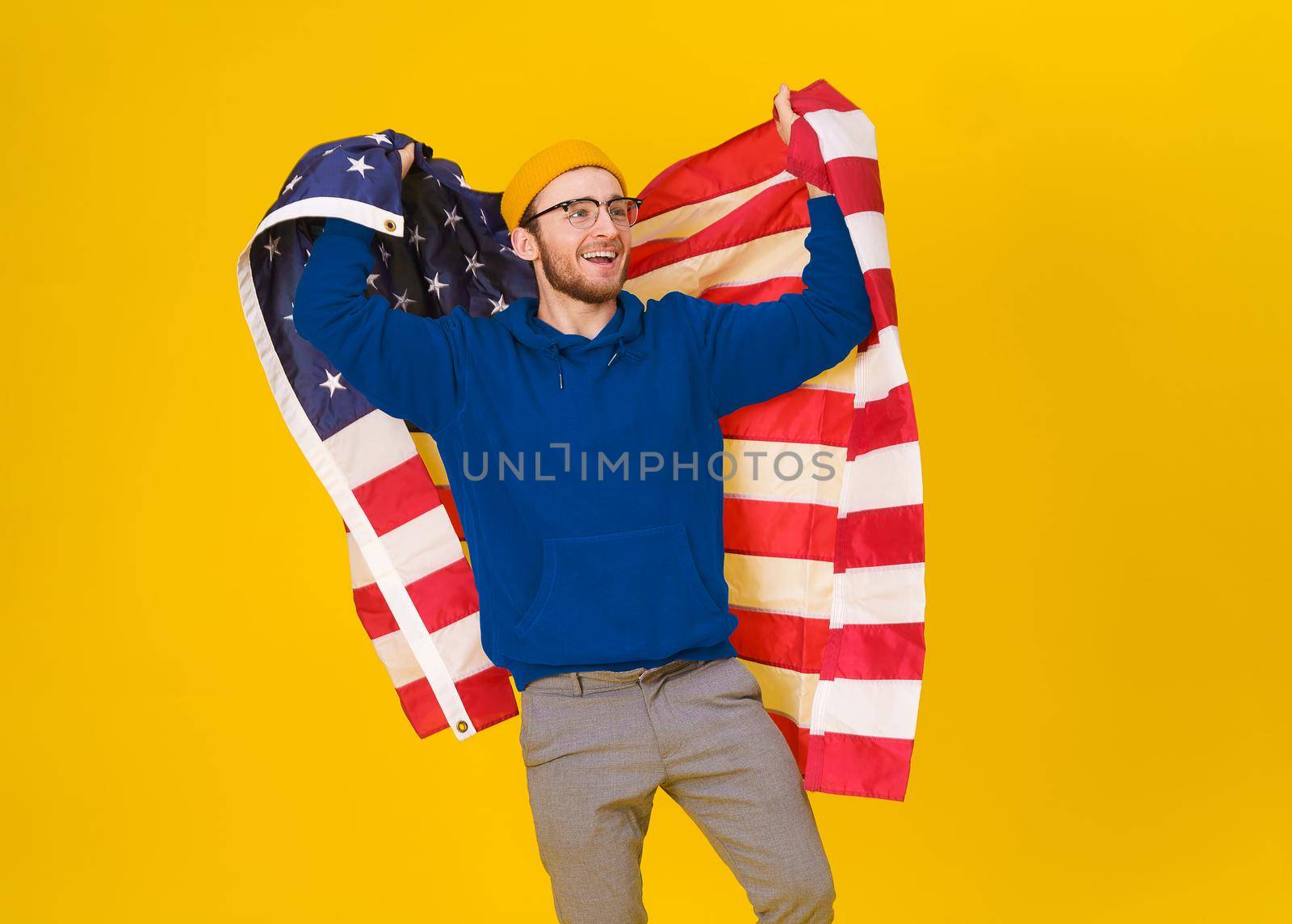 Handsome caucasian young man celebrates 4th of july in blue hoodie and USA flag behind his back isolated on yellow background. Freedom is in your life. Cheerful young man with American flag.