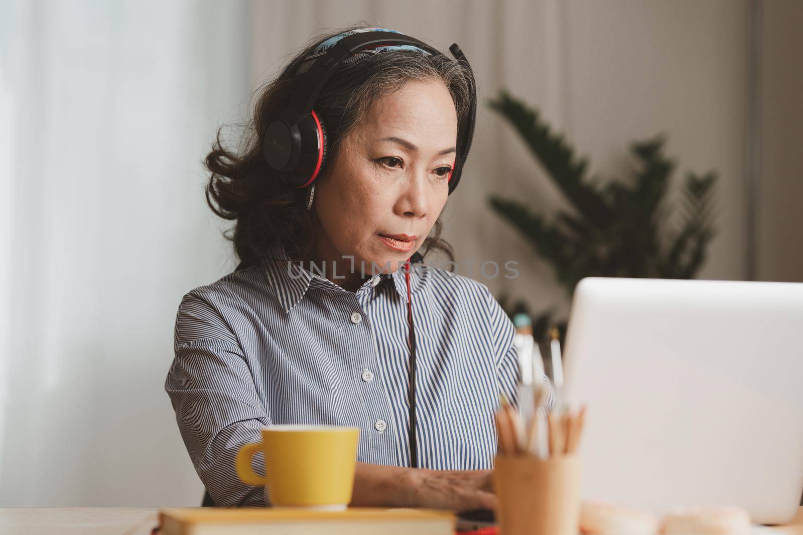 Portrait of Asian Senior Business woman wear headphone and with coffee using video call at morning at home. Account and Finance concept