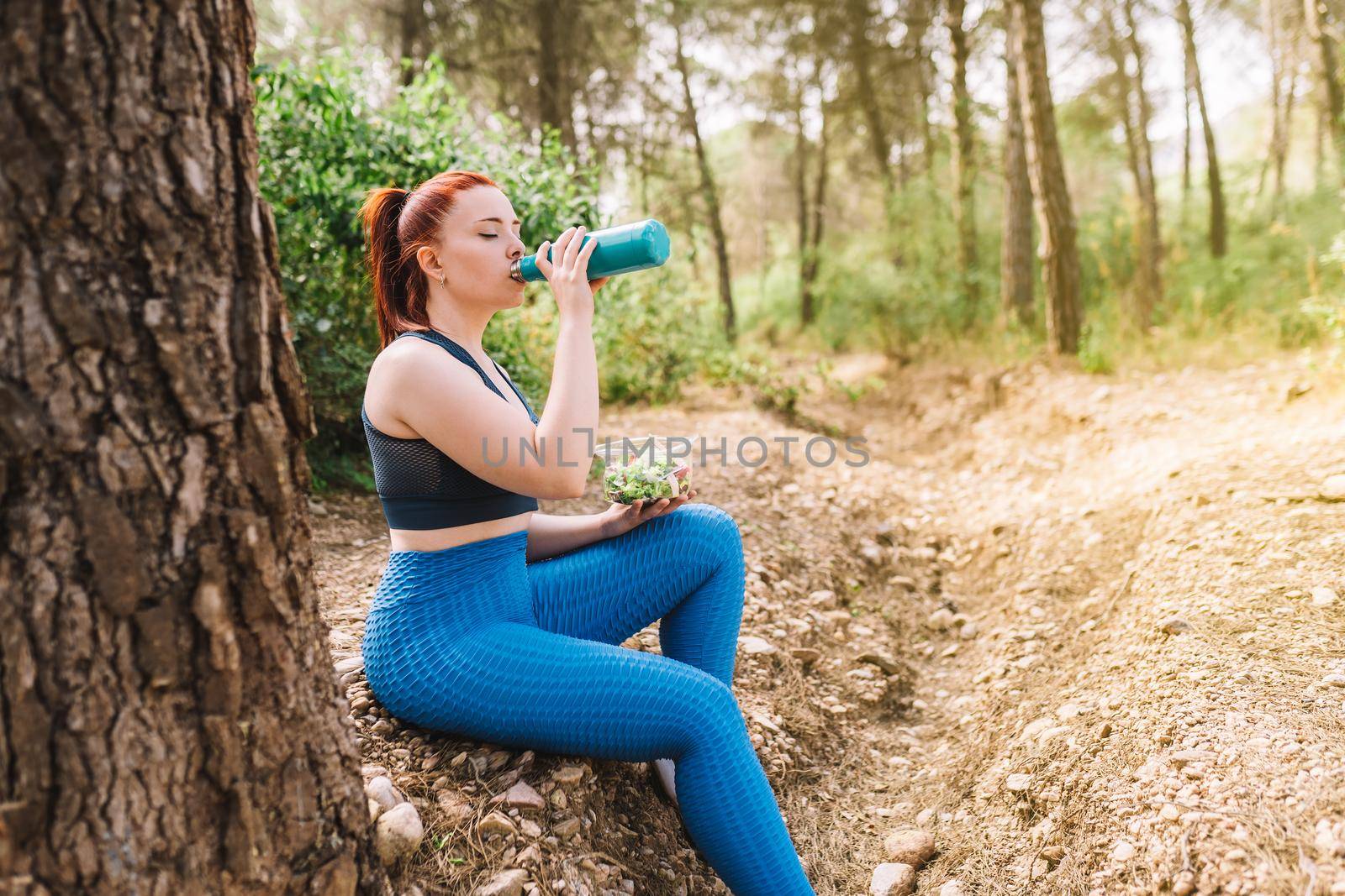 side image of a young fitness girl sitting drinking water to refresh herself from a bottle after a sports workout. girl practising sport outdoors. health and wellness lifestyle. outdoor public park, natural sunlight.