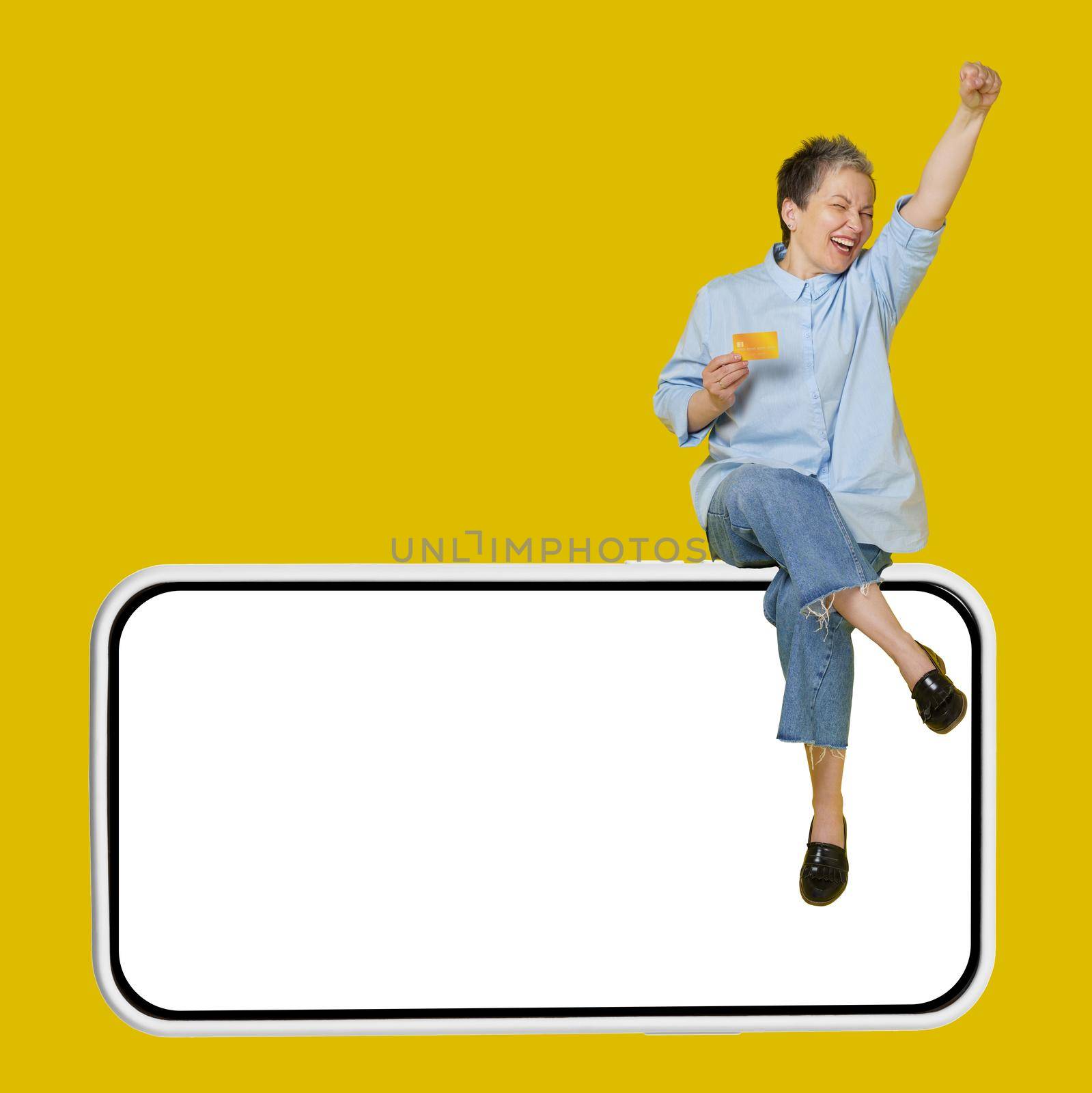 Excited, happy mature grey haired woman sitting on a giant, huge smartphone, in casual blue shirt and jeans with hand raised up in a win, isolated on yellow background.