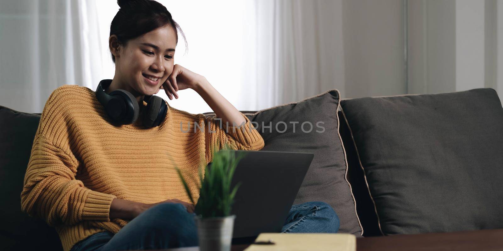 Cheerful freelancer working on laptop sitting on couch and listening to music in headphones, copyspace by wichayada