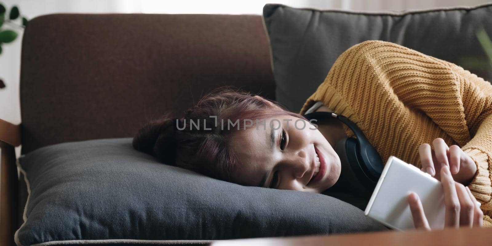 Lazy young woman spend weekend at home lying down on couch wearing headphones looking at smart phone screen listening music online. by wichayada